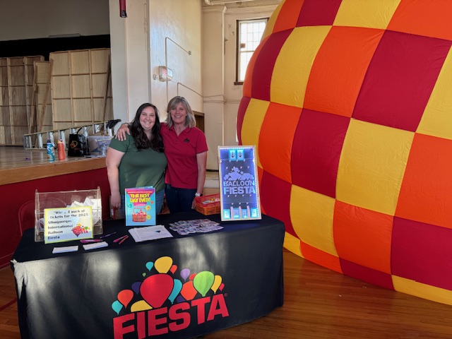 Jory and Tracey Two people stand smiling beside a colorful booth with "Fiesta" logo, inside a hall with a large checkered balloon backdrop.