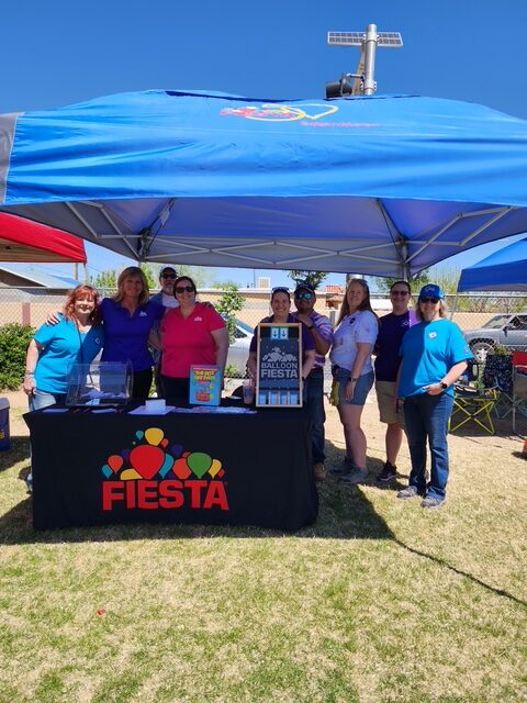 La Mesa Elem Group Several people stand under a blue canopy at a Fiesta event, with a table displaying promotional materials on grass. Bright, sunny weather.