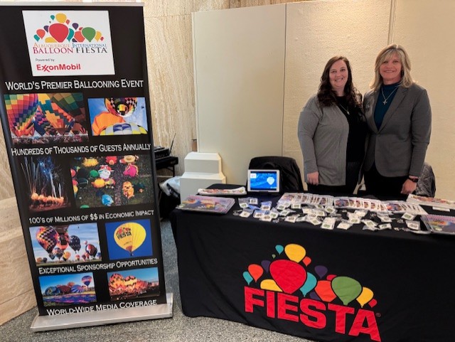 Outreach Two people stand at a promotional booth for the Albuquerque International Balloon Fiesta, featuring colorful hot air balloons and informational displays.