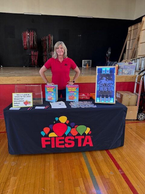 Tracey A person stands behind a Fiesta-themed table in a gymnasium, promoting balloon-related items. Various promotional materials are displayed.