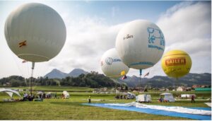 Hot air balloons prepare for launch in a grassy field with mountains in the background. People gather under tents, enjoying the event.