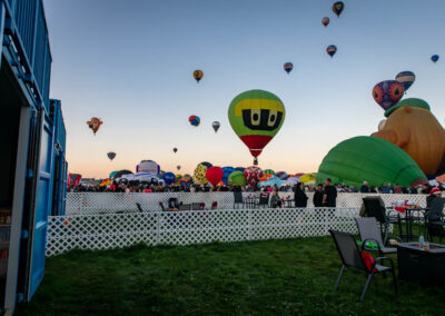 A colorful hot air balloon festival with various shaped balloons in the sky, numerous people observing, and seating areas surrounded by white fencing.