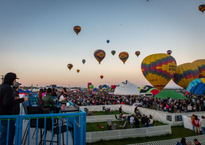 Colorful hot air balloons float above a large crowd at a festival, with people watching from a platform. No landmarks identified.