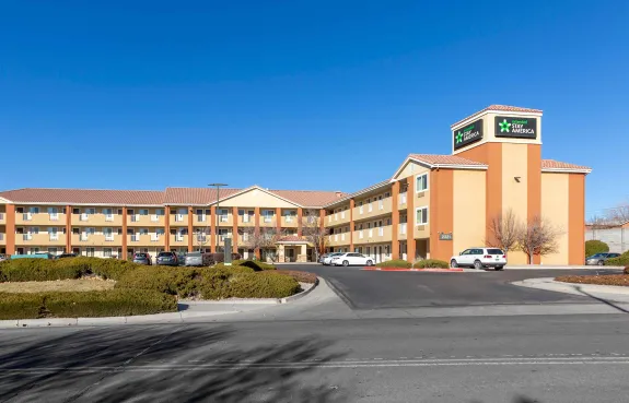 Tall hotel building with multiple windows, surrounded by trees and parked cars. Mountains are visible in the background under a clear blue sky.