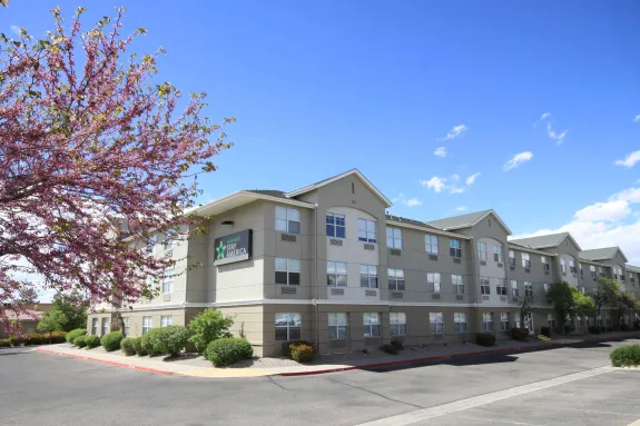 Tall hotel building with multiple windows, surrounded by trees and parked cars. Mountains are visible in the background under a clear blue sky.
