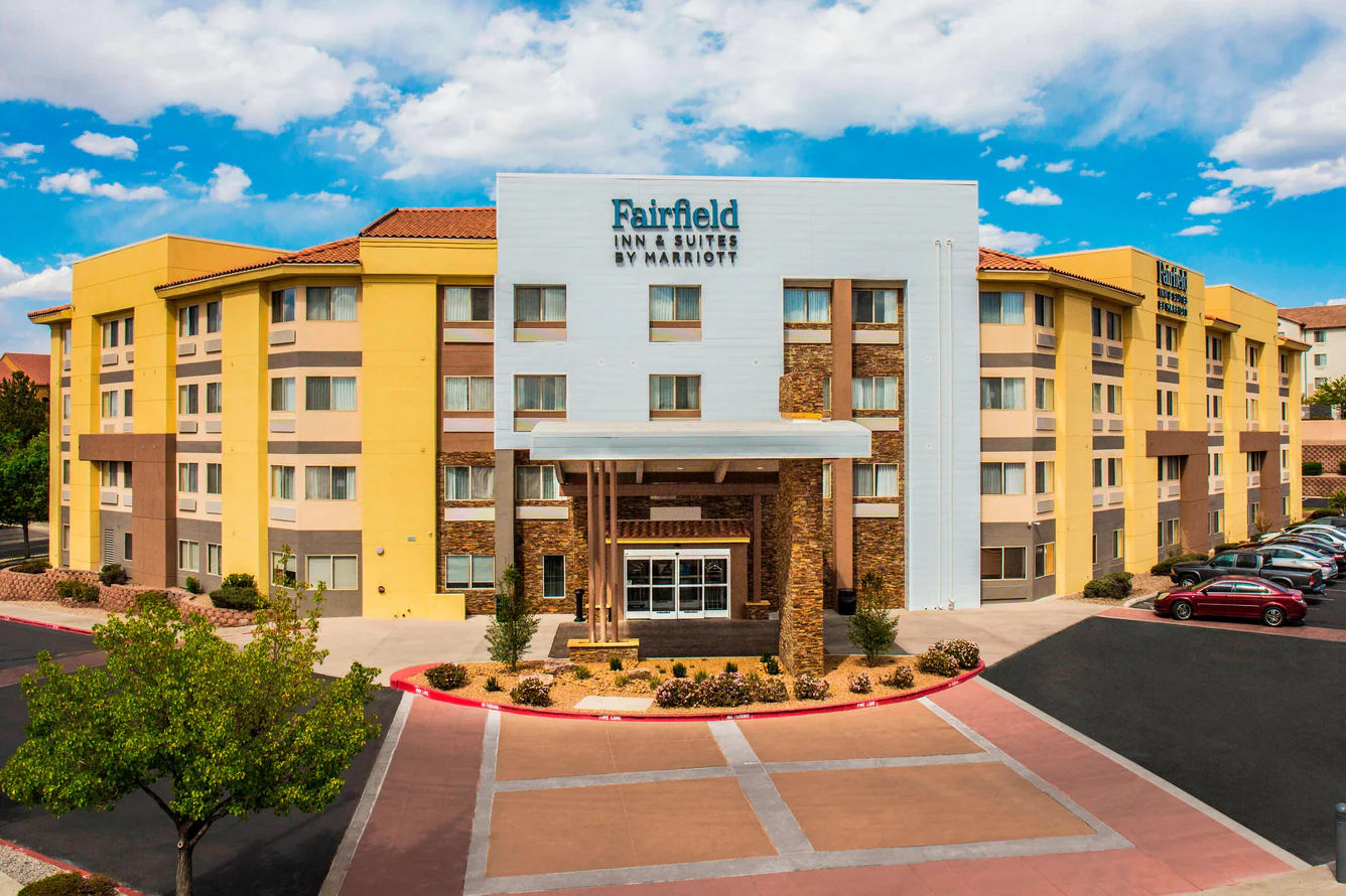 Tall hotel building with multiple windows, surrounded by trees and parked cars. Mountains are visible in the background under a clear blue sky.