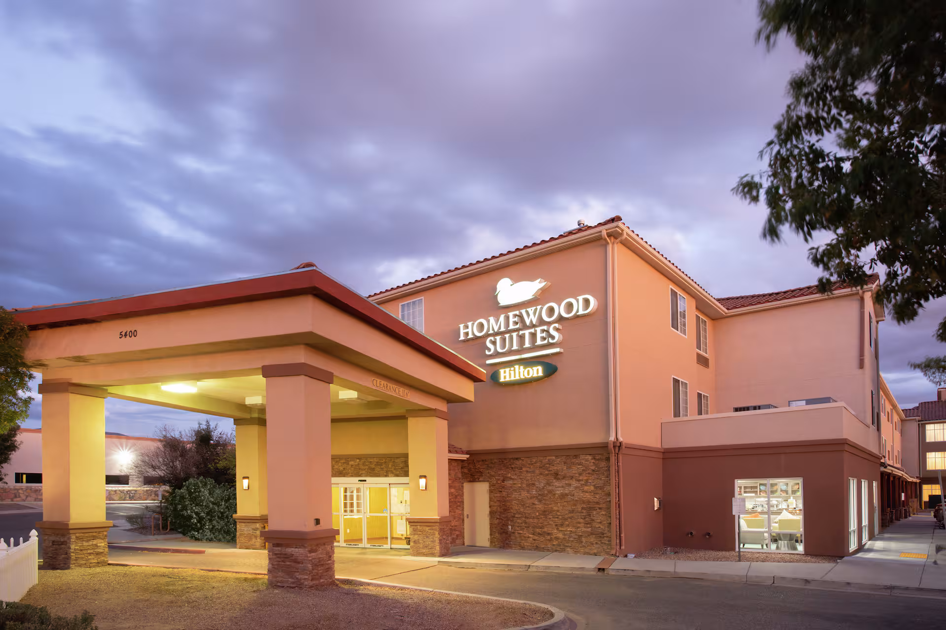 Tall hotel building with multiple windows, surrounded by trees and parked cars. Mountains are visible in the background under a clear blue sky.