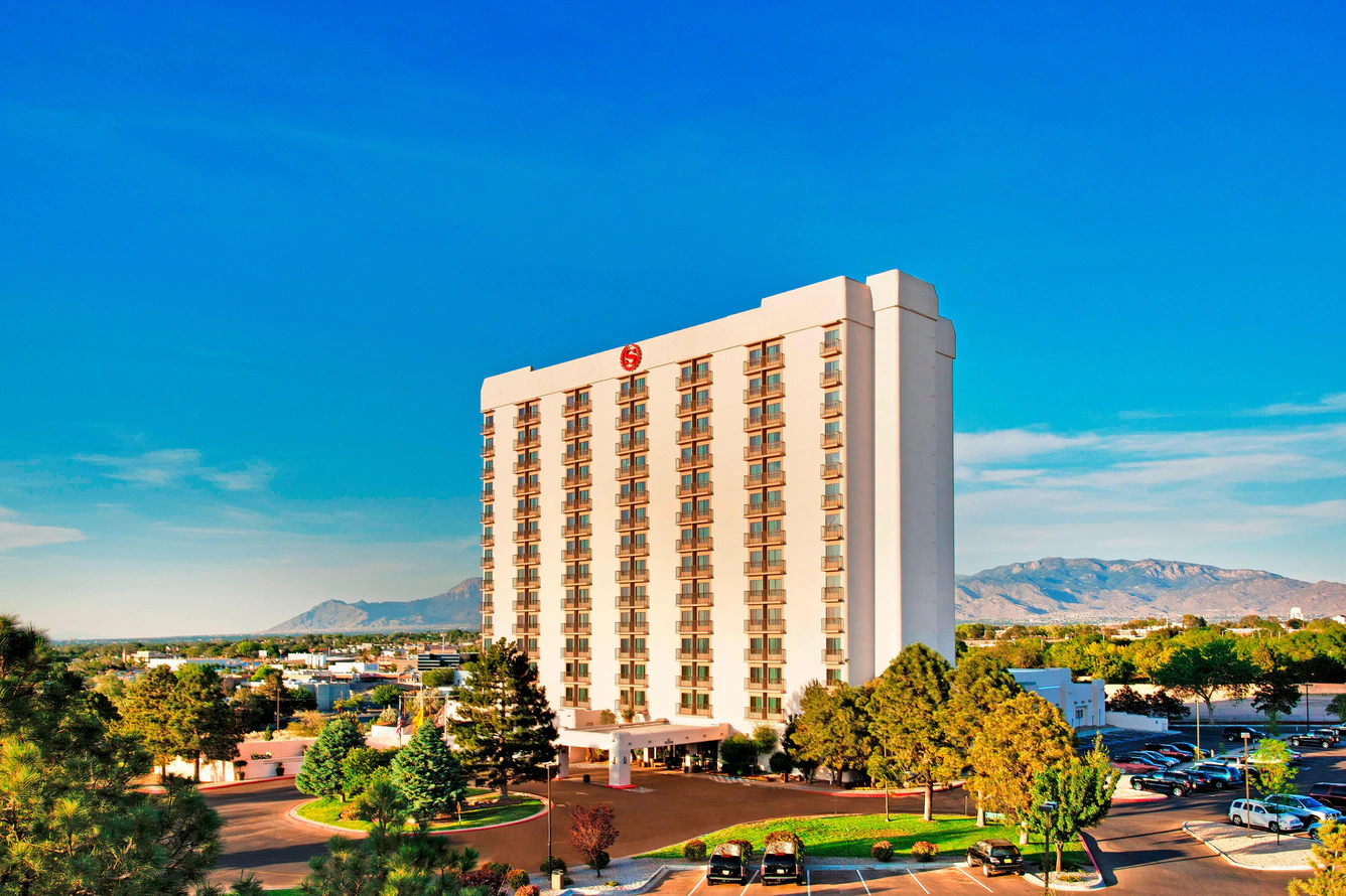 Tall hotel building with multiple windows, surrounded by trees and parked cars. Mountains are visible in the background under a clear blue sky.