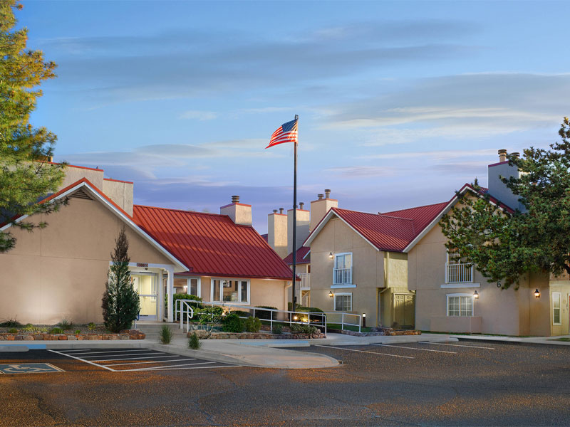 Tall hotel building with multiple windows, surrounded by trees and parked cars. Mountains are visible in the background under a clear blue sky.