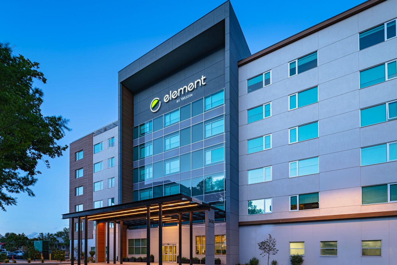 Tall hotel building with multiple windows, surrounded by trees and parked cars. Mountains are visible in the background under a clear blue sky.