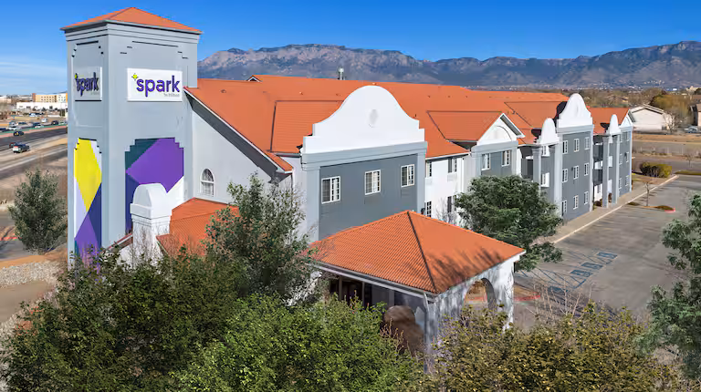 Tall hotel building with multiple windows, surrounded by trees and parked cars. Mountains are visible in the background under a clear blue sky.