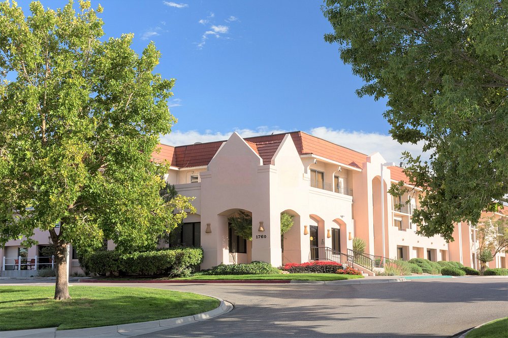 Tall hotel building with multiple windows, surrounded by trees and parked cars. Mountains are visible in the background under a clear blue sky.