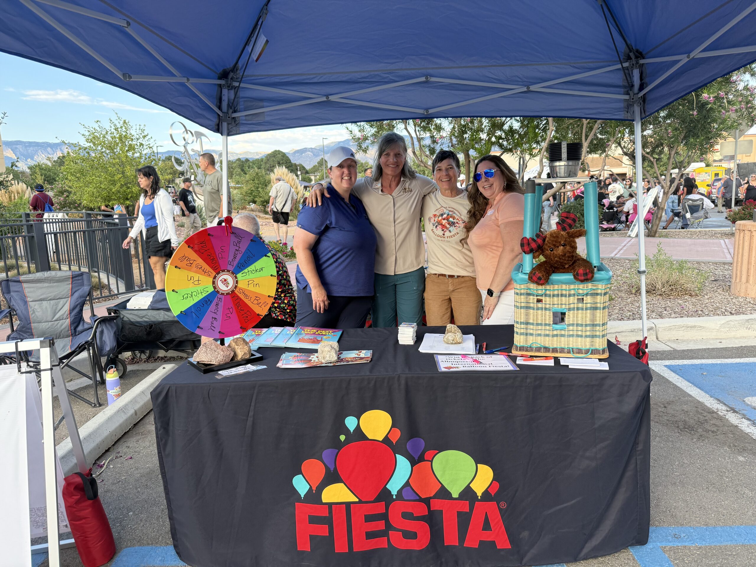 Winrock 6 Two people stand smiling beside a colorful booth with "Fiesta" logo, inside a hall with a large checkered balloon backdrop.