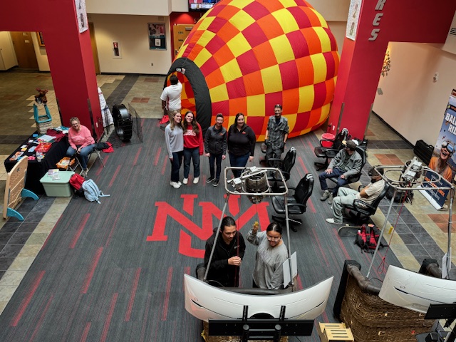 unm6 A person stands behind a Fiesta-themed table in a gymnasium, promoting balloon-related items. Various promotional materials are displayed.