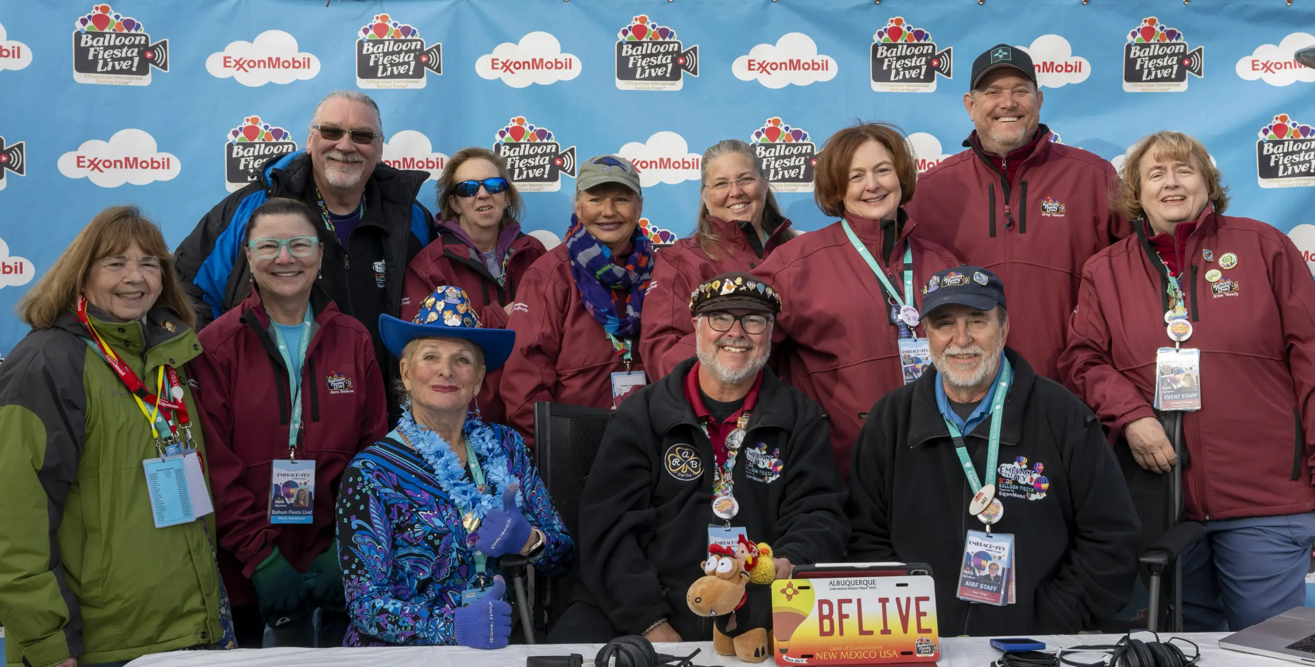 2024 BFLIveTeam Group of cheerful people in colorful attire posing together at a Balloon Fiesta event, with promotional backdrop featuring logos and badges.