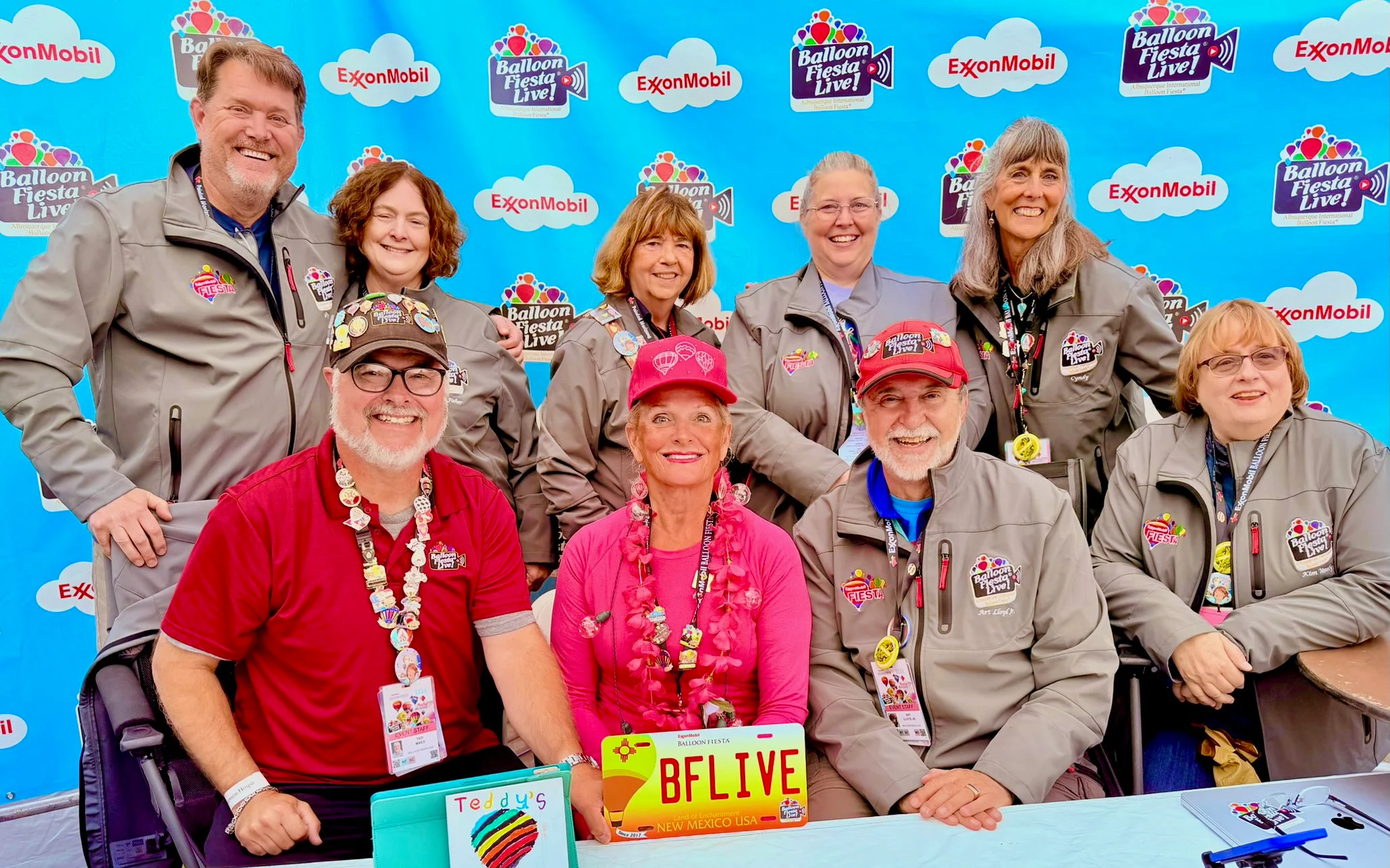BFLiveTeam2025 A group photo of nine people at a Balloon Fiesta event, wearing branded jackets and lanyards, against a bright, logo-filled backdrop.