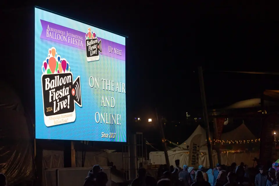 balloon-fiesta-live-2024-display A large electronic billboard displays "Balloon Fiesta Live!" at night, surrounded by a crowd under festive lights. The event is Albuquerque International Balloon Fiesta.