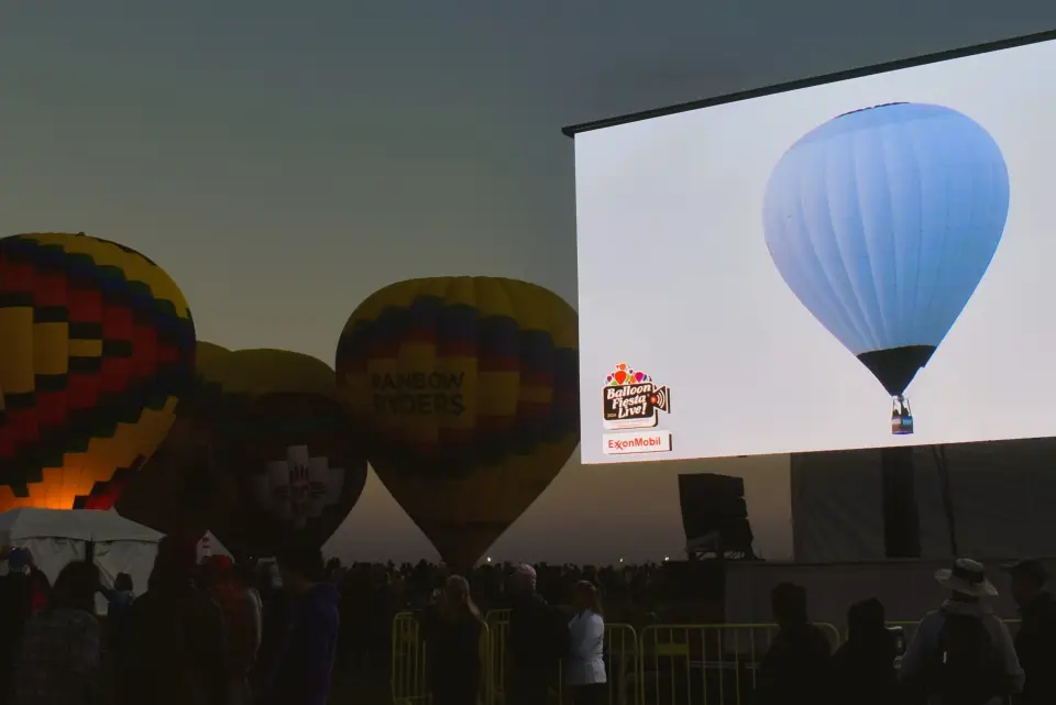 balloon-fiesta-live-broadcast A hot air balloon festival at dusk, featuring colorful balloons and a large screen displaying another balloon. Crowds gather, enjoying the event.