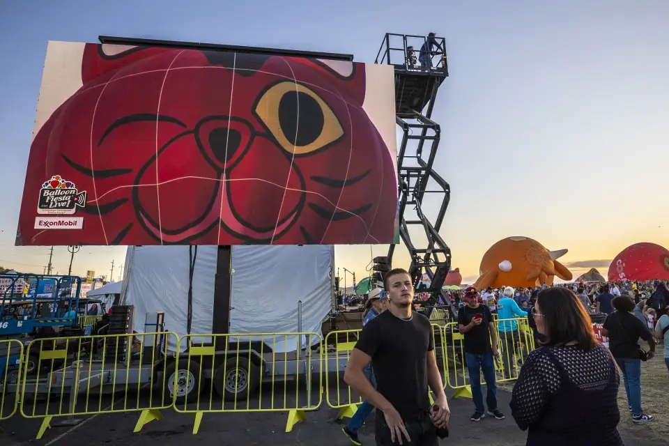 balloon-fiesta-live-broadcast02 A large crowd gathers at a balloon festival, featuring a big red cat design on a screen, surrounded by various inflatable shapes.