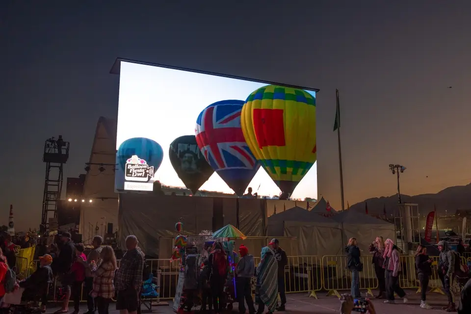 balloon-fiesta-live-broadcast03 A large screen displays colorful hot air balloons at a festival. People gather in front, surrounded by event structures and lighting.