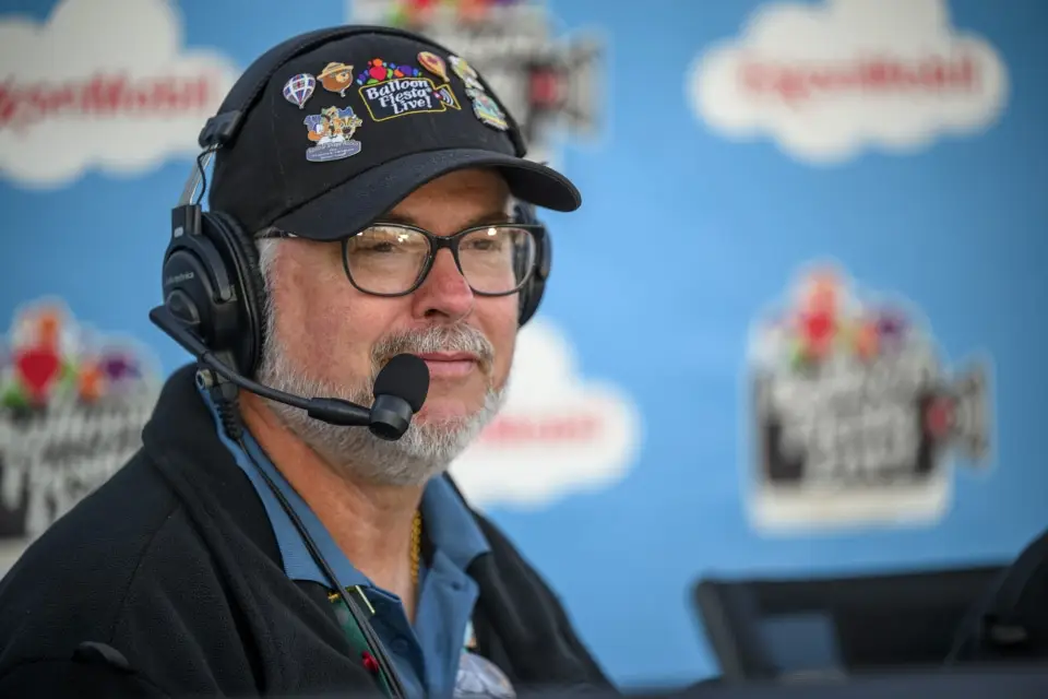 balloon-fiesta-live-broadcaster Balloon Fiesta Live broadcaster wearing a headset and cap decorated with pins. Background has logos and cloud designs on a blue backdrop.