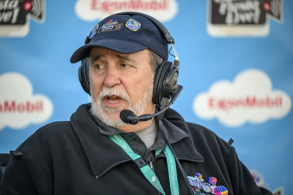 balloon-fiesta-live-broadcaster02 Balloon Fiesta Live broadcaster with a headset speaks into a microphone. They wear a cap and jacket with colorful patches. Blue backdrop displays "ExxonMobil" logo.