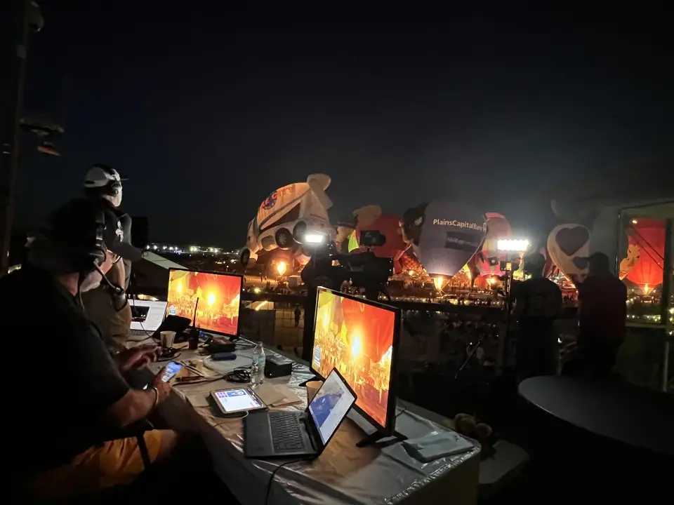 balloon-fiesta-live-broadcasters-glow Balloon Fiesta Live crew members work at computers during a nighttime hot air balloon event with glowing displays, under bright lights, in an outdoor setting.
