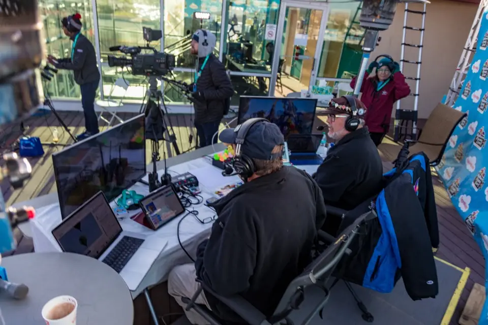 balloon-fiesta-live-broadcasters-in-studio Balloon Fiesta Live broadcasters with cameras, monitors, and laptops are setting up a broadcast studio outside a glass building. Equipment and colorful signs are visible.