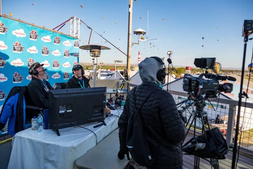 balloon-fiesta-live-broadcasters Balloon Fiesta Live broadcasters at a table with cameras, surrounded by hot air balloons in the sky during an outdoor event.