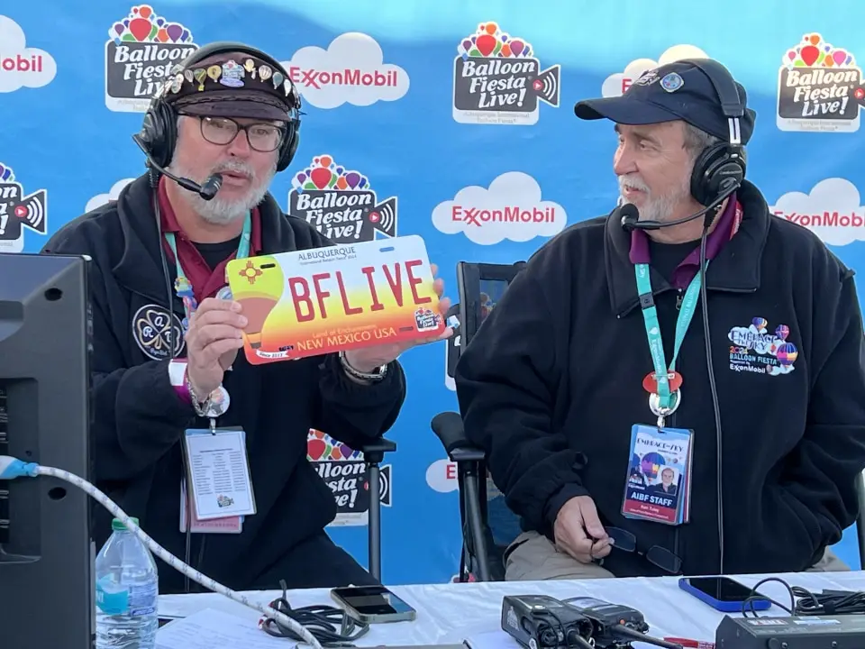balloon-fiesta-live-broadcasters02 Balloon Fiesta Live broadcasters with headsets discuss the Albuquerque Balloon Fiesta, featuring a custom license plate, at a broadcast booth with promotional branding in the background.