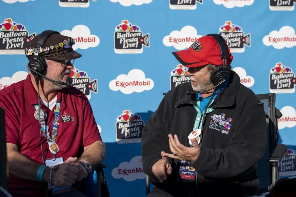 balloon-fiesta-live-broadcasters04 Balloon Fiesta Live broadcasters wearing headsets converse at Balloon Fiesta Live event. They're seated against a backdrop with logos, including ExxonMobil.