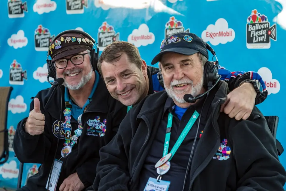 balloon-fiesta-live-broadcasters05 Three people wearing headsets, smiling, posing together in front of a Balloon Fiesta Live backdrop with logos, including ExxonMobil.