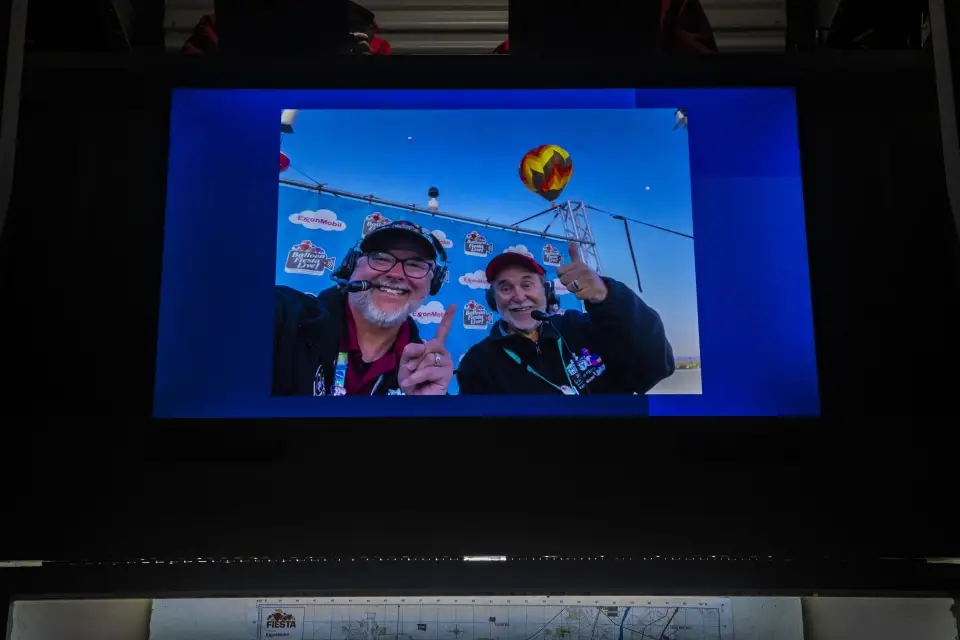 balloon-fiesta-live-broadcasters06 Balloon Fiesta Live broadcasters smiling in a selfie on a display screen, with a hot air balloon and logos in the background.