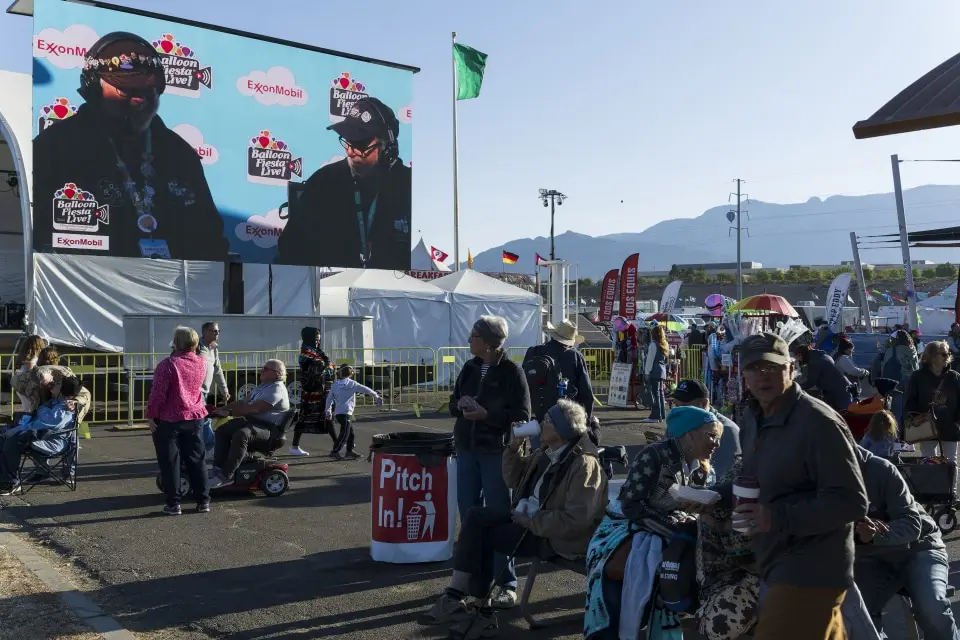 balloon-fiesta-live-broadcasters07 People gather outdoors at Balloon Fiesta with a large screen displaying Balloon Fiesta Live broadcasters. Flags and mountains are visible in the background.