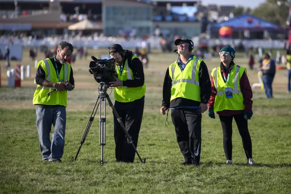 balloon-fiesta-live-camera-operators-on-field Four Balloon Fiesta Live crew members in bright vests operate camera equipment on a grassy field, focused on a distant event. Buildings and spectators in the background.
