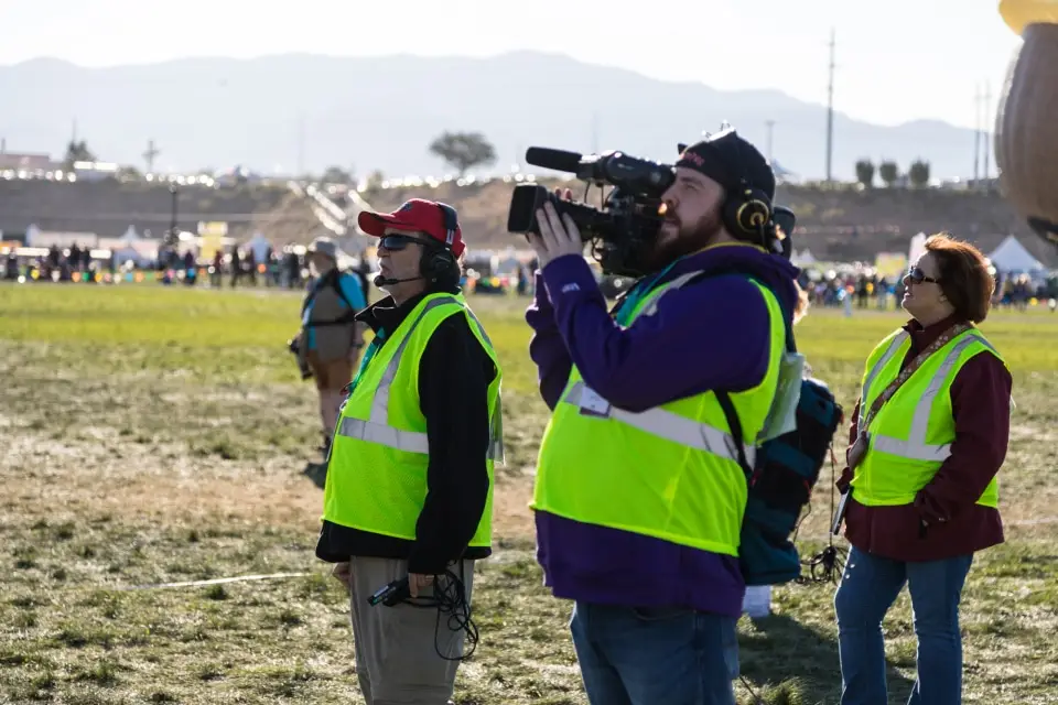 balloon-fiesta-live-camera-operators-on-field02 Three Balloon Fiesta Live crew members wearing safety vests stand on grass; one films with a large camera. A crowd is visible in the background.