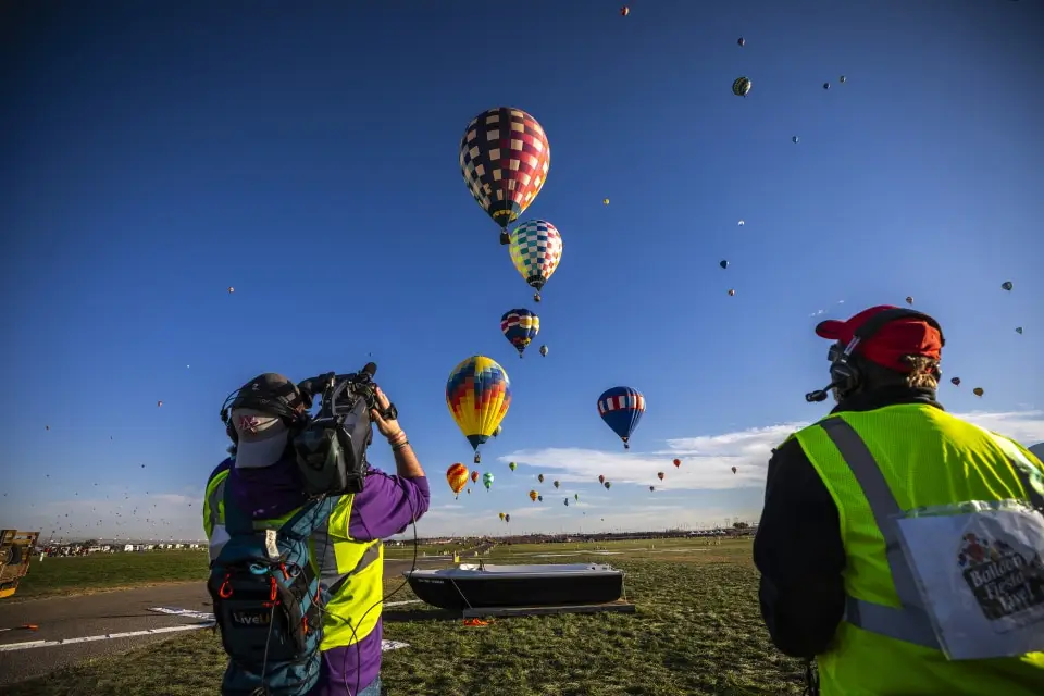 balloon-fiesta-live-camera-operators-on-field03 Two Balloon Fiesta Live crew members in yellow vests watch and film colorful hot air balloons ascending into a clear blue sky over a grassy field.