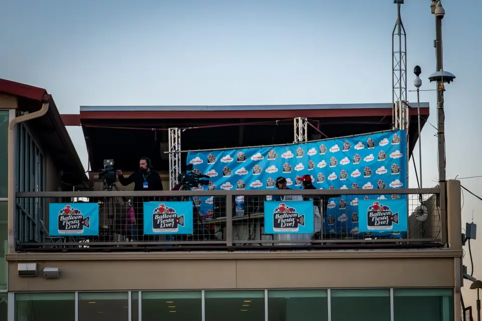 balloon-fiesta-live-camera-operators A person operates a camera on a rooftop set with "Balloon Fiesta Live" banners, surrounded by technical equipment and a clear sky backdrop.