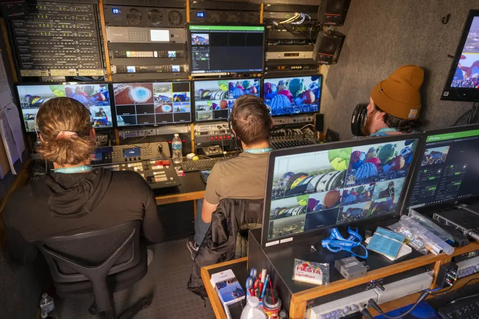 balloon-fiesta-live-control-room03 Three people operate video equipment inside the Balloon Fiesta Live control room filled with screens displaying vibrant balloon visuals and technical controls