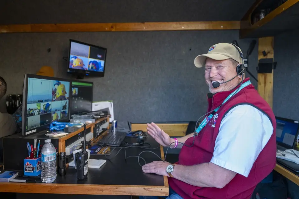 balloon-fiesta-live-control-room04 A person wearing a headset sits at a Balloon Fiesta Live workstation with multiple screens in a control room setting, surrounded by electronic equipment and monitors.