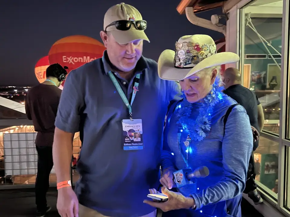 balloon-fiesta-live-crew-glow Two people converse during the balloon glow. One wears a light-up blue outfit. A hot air balloon is visible in the background.