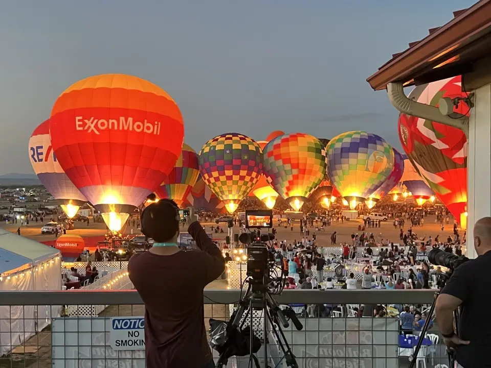 balloon-fiesta-live-crew-taking-photo The Balloon Fiesta grounds are packed with people watching the balloon glow. Camera equipment is set up for capturing the vibrant scene.