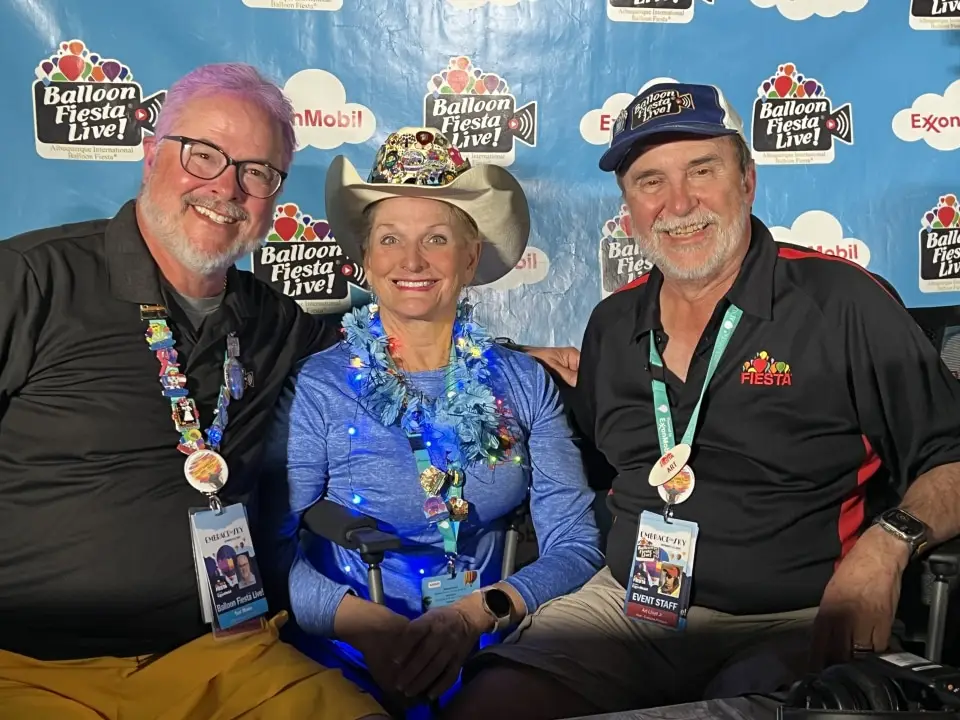 balloon-fiesta-live-crew Three people smiling at Balloon Fiesta Live booth, wearing branded lanyards and festive accessories, against a backdrop with logos and sponsor names.