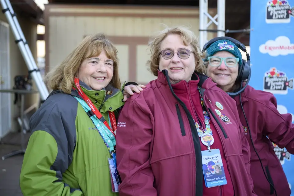 balloon-fiesta-live-crew02 Three people in jackets pose for a cheerful group photo Balloon Fiesta Live studio, with badges and lanyards visible, against a simple backdrop.