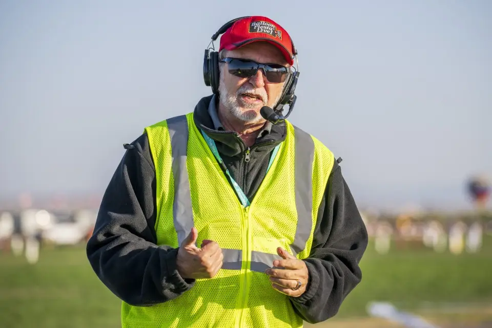 balloon-fiesta-live-crewperson-on-field Balloon Fiesta Live crew member wearing a headset and yellow safety vest stands outdoors, gesturing. They are likely involved in event coordination or management.
