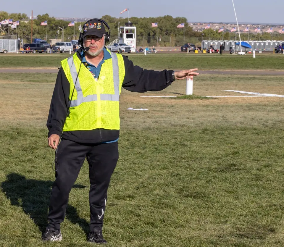 balloon-fiesta-live-crewperson-on-field02 Balloon Fiesta Live crew member in a safety vest and headset gestures on a grassy field with scattered flags and parked vehicles in the background.
