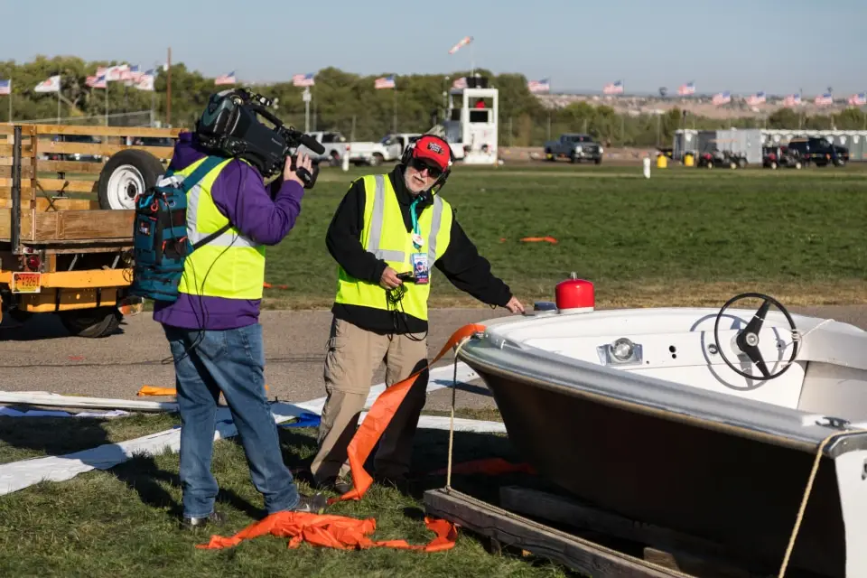 balloon-fiesta-live-crewperson-on-field03 Two people in safety vests next to a boat shaped hot air balloon basket on grass, camera filming, flags in background. Event setup environment.