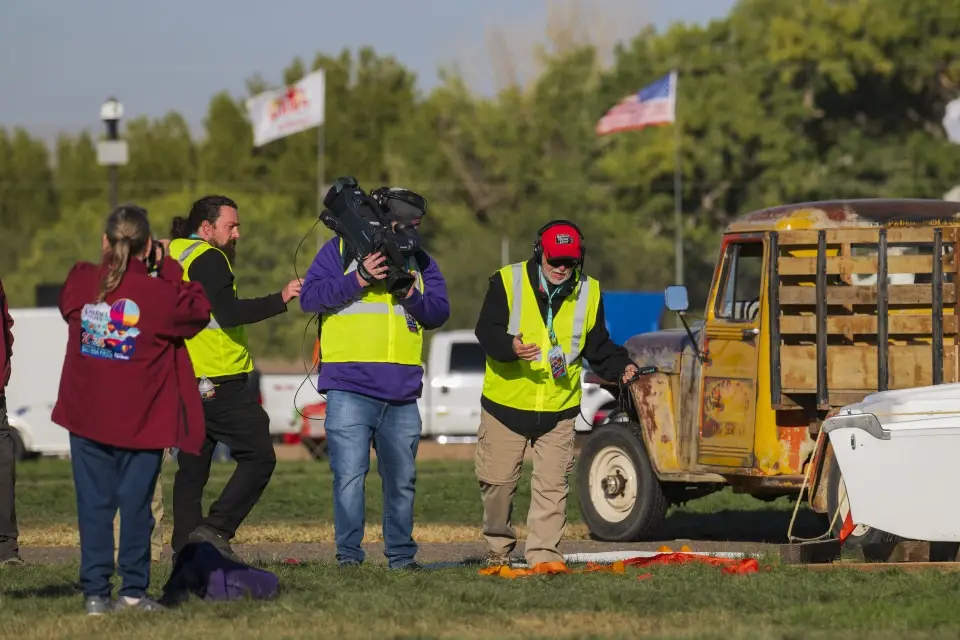 balloon-fiesta-live-crewperson-on-field04 A group of people in safety vests interacts near a vintage truck on Balloon Fiesta grounds, with flags and trees in the background.