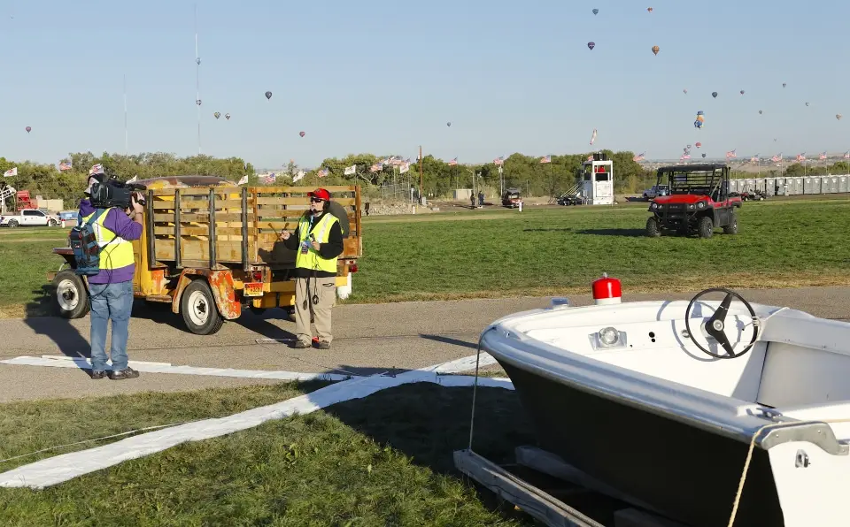 balloon-fiesta-live-crewperson-on-field05 Two people, one with a camera, near a trailer and a boat shaped hot air balloon basket. Hot air balloons fill the sky