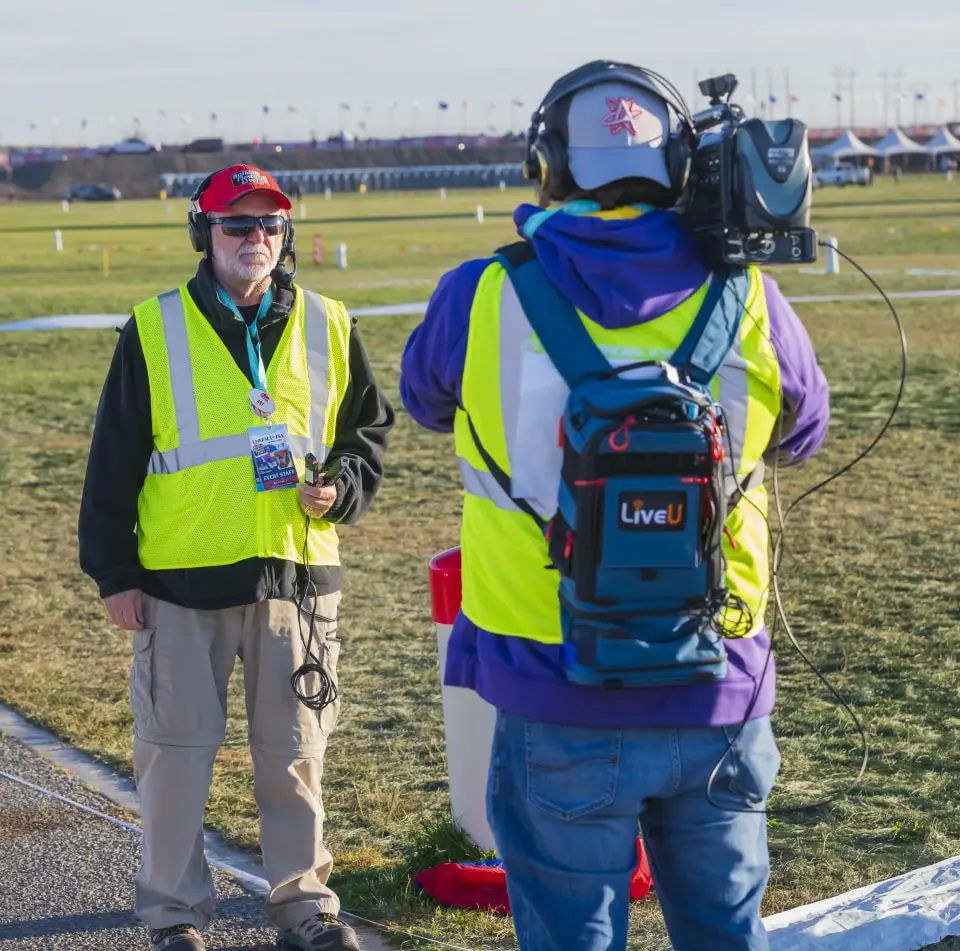 balloon-fiesta-live-crewperson-on-field06 Two persons in neon vests, with broadcasting equipment, stand outdoors on grassy field, preparing for a live event.
